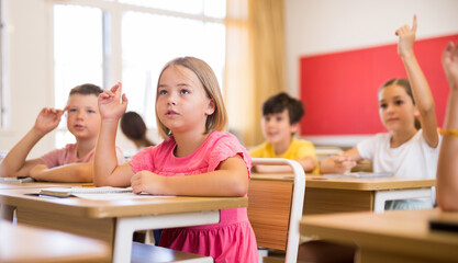 Portrait of positive pupil girl raising hand to answer during lesson at classroom