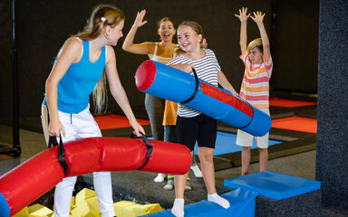 Emotional active preteen girl having funny wrestling by pugil sticks on battle beam with other girl...