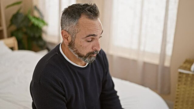 A mature hispanic man with grey beard appears contemplative in a bedroom setting.