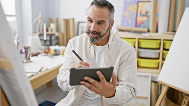 A cheerful bearded man using a tablet in a creative studio, surrounded by art supplies and paintings.