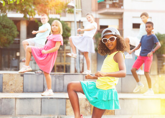 Confident african american preteen b-girl in sunglasses and cap dancing with group of friends on...