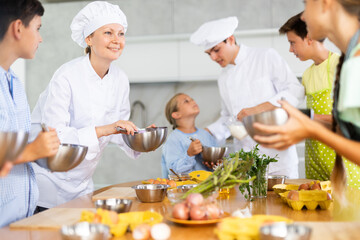 Positive woman professional chef in white uniform conducting culinary class, imparting cooking skills to group of interested tween boys and girls