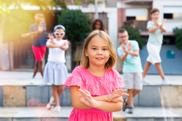 Positive cute girl dancing modern choreography with group of tweenagers on city street on summer day..