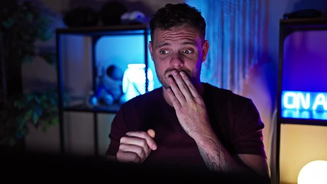 Shocked young man covering mouth, surprising expression while playing video game in gaming room. afraid for his unanticipated mistake in-game