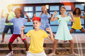 Positive modern tween boy with dyed red hair dancing with group of friends in sunbeams on summer...