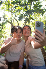 Asian Thai daughter and elder mother taking selfie together with happy smiling, Mom show thumbs up, both spending time together at cafe in garden on holidays.