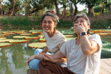 Side view of asian Thai Chinese elder mother and daughter sitting on the bank by lotus flower lake...