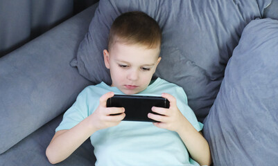 Child is focused on playing on his smartphone while sitting on couch at home. Problem with online game addiction. Spending time at home, leisure, relaxation, happy carefree childhood