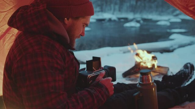 Photographer sitting in tent and taking pictures of bonfire with professional camera when having camping trip in winter