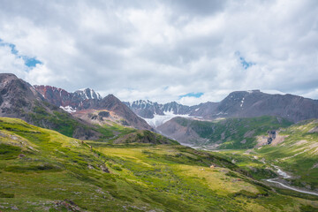 Colorful vast scenery of wide green alpine valley with panoramic view to large snow-capped top, sharp rockies, rocky pointy peak, snowy mountain range and big glacier tongue far away under cloudy sky.