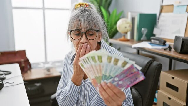 Grey-haired, middle age woman shocked, surprised and afraid at work! hand covering mouth in silent shock holding hong kong banknotes, an office mistake causing fear, shame and mute surprise.