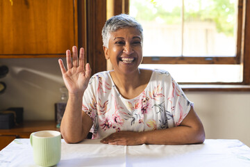 A mature biracial woman greets warmly from her home kitchen on a video call