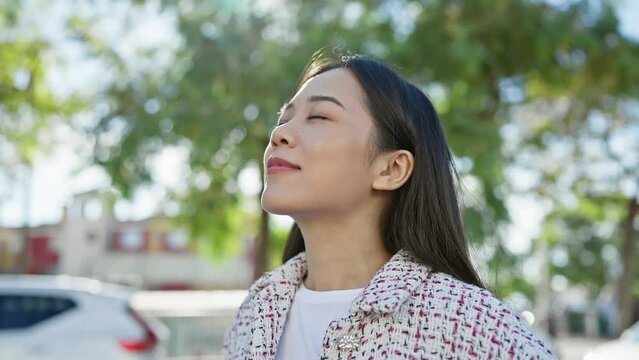 A serene young asian woman enjoys a tranquil moment outdoors on a sunny city street.