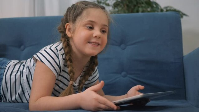 Little kid girl using tablet device, lying on sofa. Smiling little girl watching online cartoons on tablet on Internet using digital tablet technology device. kid plays at home sofa computer addiction