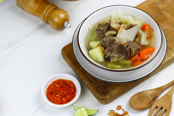 Sop daging sapi or Indonesian beef soup, served on ceramic white bowl with sambal and slices of lime. Selective focus with white marble background. 