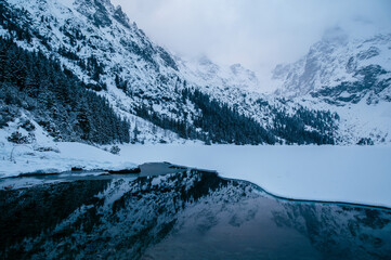 Obraz premium Morskie Oko lake sits in front of a majestic snowy mountain range, creating a stunning natural landscape. The sky is filled with fluffy clouds, enhancing the atmosphere of the scene. Tatry, Poland