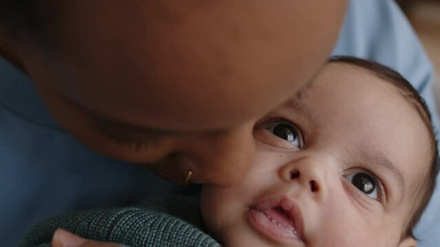Closeup of happy young African American woman kissing little baby son on cheek while spending daytime together at home