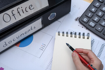 Dokumenty księgowe w segregatorach leżą na biurku w firmie obok kalkulatora, kobieta oblicza koszty 