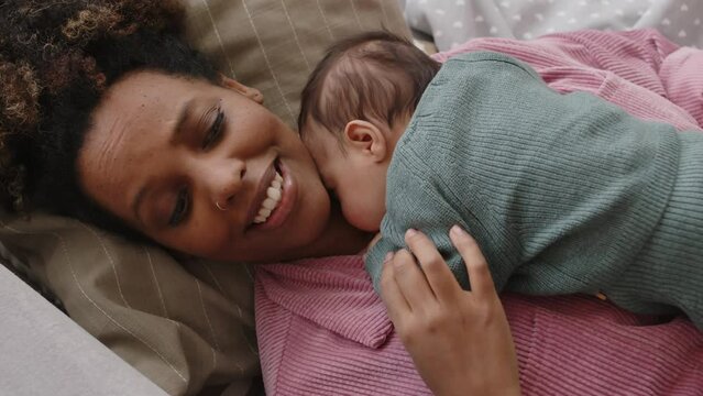 Top-view shot of young happy Black woman lying on sofa with little baby on her chest
