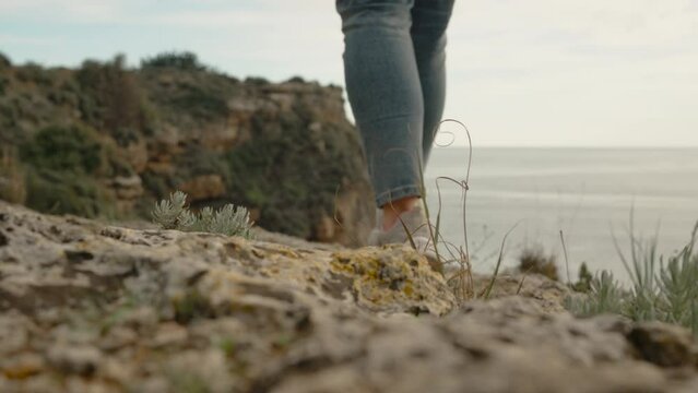 Tourist Woman Steps on Rocks and Takes Off Her Backpack at the Edge of a Cliff by the Sea. View from Below, Close-Up, the Focus Follows the Feet.