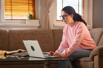 Young Caucasian woman wearing glasses is sitting on couch and using her laptop. Cozy interior....