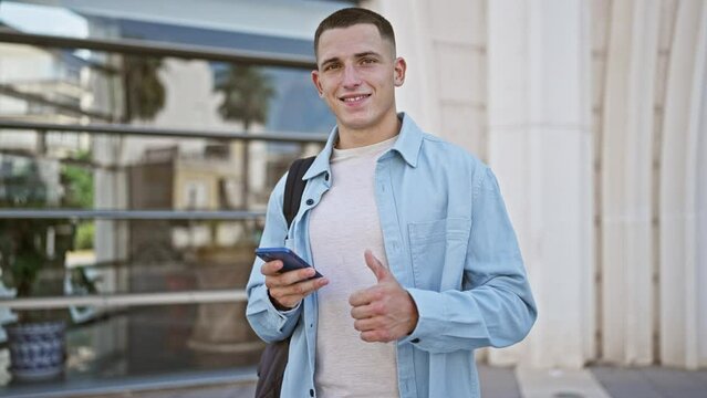 A young adult man in casual attire giving a thumbs up while holding a smartphone in an urban setting.