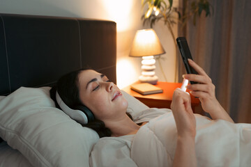 Young Caucasian woman wearing headphones is lying in bed at night and using smartphone, listening...