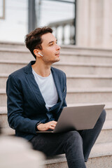 Dreaming young man wearing ear-pods works on laptop while sitting outdoors. Vertical shot.