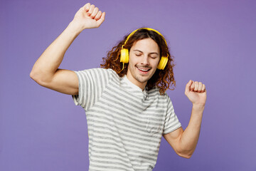 Young fun joyful man he wear grey striped t-shirt casual clothes listen to music in headphones raise up hands dance isolated on plain pastel light purple background studio portrait. Lifestyle concept