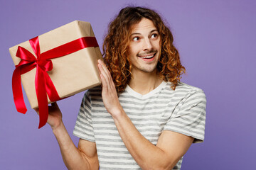 Young man wear grey striped t-shirt casual clothes hold in hand present box with gift ribbon bow look aside on area isolated on plain pastel light purple background studio portrait. Lifestyle concept