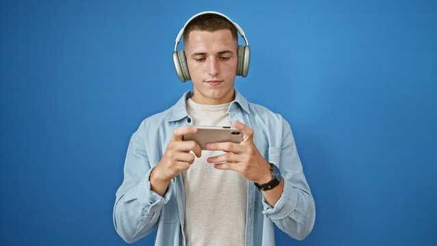 Young adult man with headphones using smartphone against blue wall, expressing engagement and concentration.