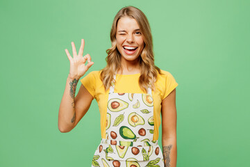 Young smiling cheerful happy fun housewife housekeeper chef cook baker woman wearing apron yellow t-shirt look camera wink eye isolated on plaint pastel green background studio. Cooking food concept.