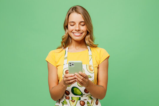 Young housewife housekeeper chef cook baker woman wear apron yellow t-shirt hold in hand use mobile cell phone look for recipe isolated on plain pastel green background studio. Cooking food concept.
