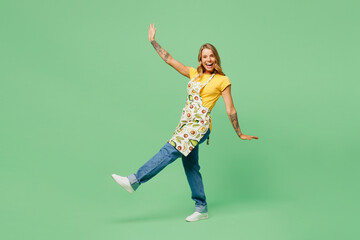 Full body side view fun young housewife housekeeper chef cook baker woman wear apron yellow t-shirt raise up leg look camera walk dance isolated on plain green background studio. Cooking food concept.