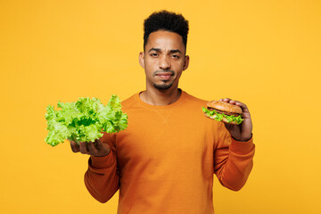 Young sad man wear orange sweatshirt casual clothes hold lettuce salad burger choose what to eat...