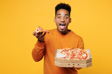 Young surprised man wear orange sweatshirt casual clothes hold eat italian pizza in cardboard...