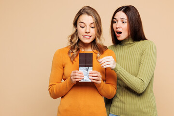 Young shocked happy friends two women they wear orange green shirt casual clothes together hold in hand chocolate bar count calories isolated on plain pastel light beige background. Lifestyle concept.