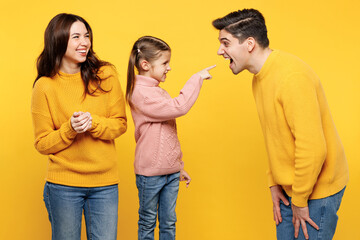 Young parents mom dad with child kid girl 7-8 years old wear pink sweater casual clothes point index finger on opened mouth of daddy play game isolated on plain yellow background. Family day concept.
