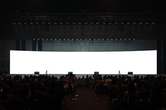 Front view of mockup on big blank white screen with space in empty huge hall with scene and silhouette of audiences