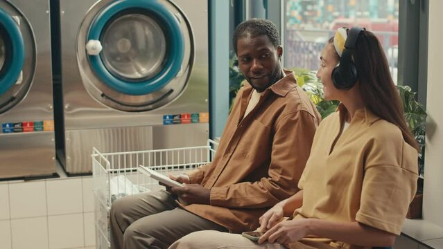 Side footage of confident Black guy with paper book making acquaintance with positive Caucasian girl with headphones at laundromat