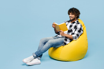 Full body smiling happy young Indian man he wears shirt white t-shirt casual clothes sit in bag chair read book isolated on plain pastel light blue cyan background studio portrait. Lifestyle concept.