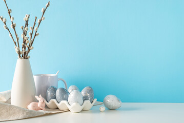 Fototapeta na wymiar Kitchen table Easter display, side view angle, showcasing a ceramic container with luxurious eggs, a rabbit, mug, and cloth, beside a vase of pussy willow, against a gentle blue wall with text space
