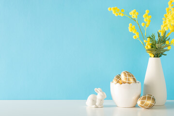 Holiday kitchen scene, showcasing a side view of a counter with a porcelain bowl of gold painted eggs, a bunny statuette, and a pitcher of tulips on a light blue wall background for text