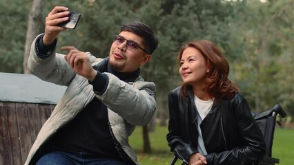 Asian man making a funny selfie video for social media with a disabled asian woman in a wheelchair on a smartphone in an autumn city park during an outdoor walk together - Powered by Adobe