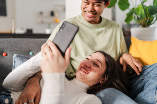 Playful and affectionate interaction as a man shares his smartphone screen with a smiling woman, intimate moment on a couch