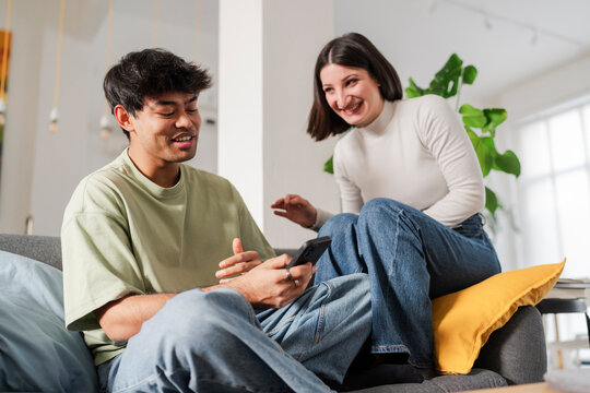 Engrossed young couple sitting on a couch, closely viewing content on a smartphone in a well-lit living room.