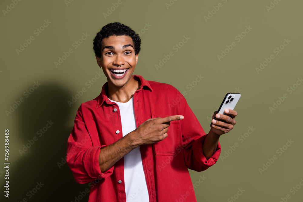 Poster photo of funky cheerful guy wear red shirt pointing finger modern gadget empty space isolated khaki 