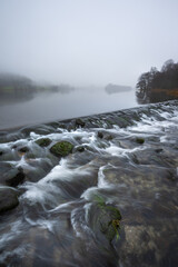 Flowing weir overlooking a misty Grasmere lake on a drizzly Winter morning in The Lake District, UK. - 736992237