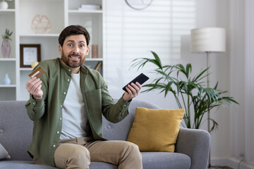 Joyful adult male with beard and casual shirt shows credit card, sitting with phone at home,...