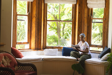 A mature biracial woman enjoys reading in a sunny home interior with copy space